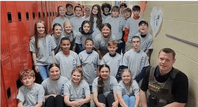 approximately 2 dozen fifth grade students in grey tshirts are crowded into a school hallyway posing with a sheriff's deputy.