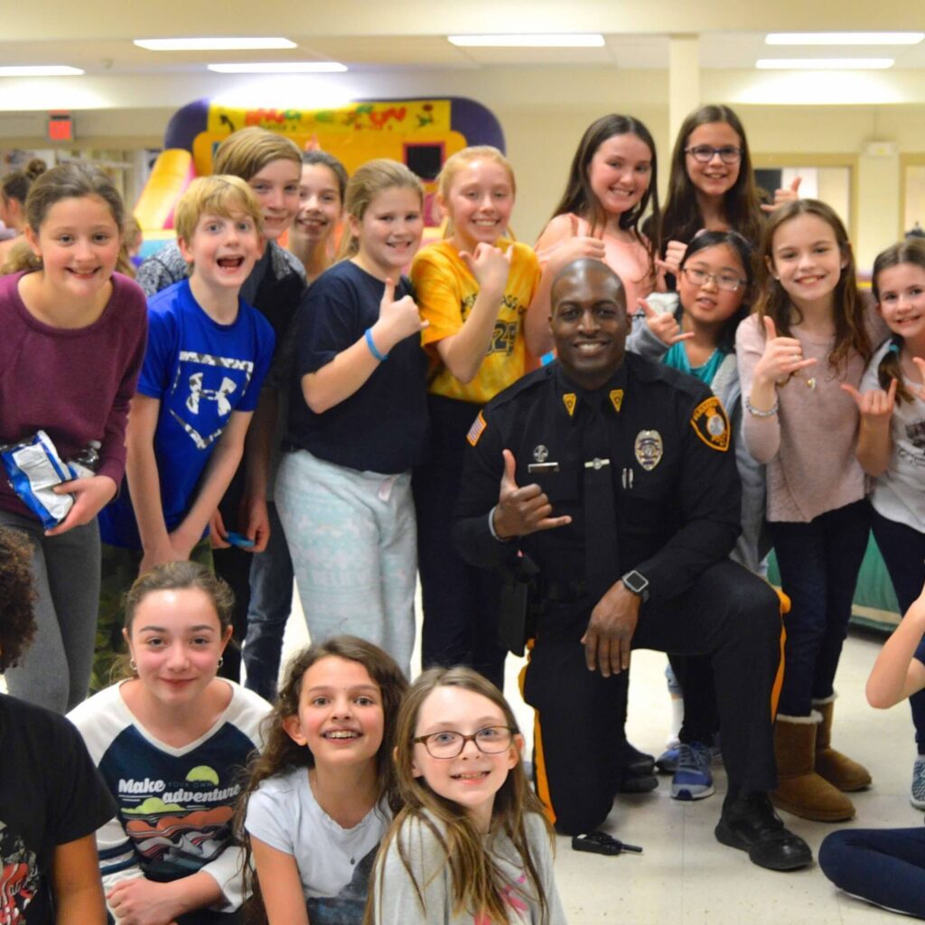 A smiling police officer is surrounded by smiling kids in a school classroom